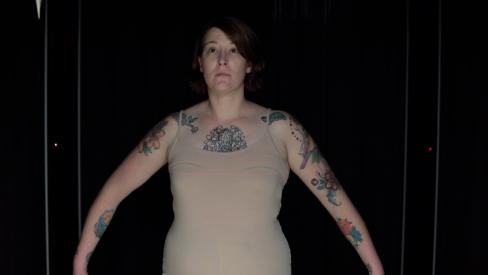 A woman in a nude body suit stands akimbo in front of a black background
